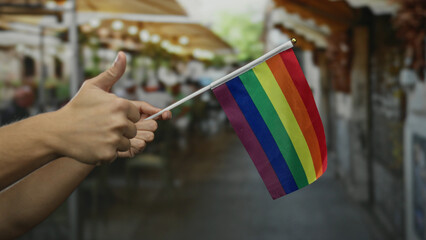 Hand holding rainbow flag with man's thumbs up in outdoor urban cafe setting showcasing support for...