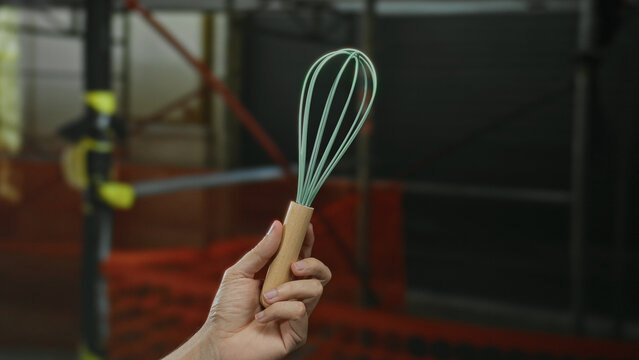 Hand holding a kitchen whisk at a construction site, highlighting a contrasting mix of culinary and industrial themes outdoors.
