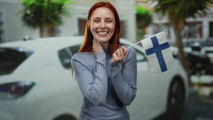 Woman smiling outdoors holding finnish flag in city street with red hair and wearing blue sweater,...