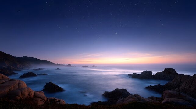 A tranquil twilight seascape with a vast starry sky over the ocean featuring rugged rocks and gentle mist along the coastline