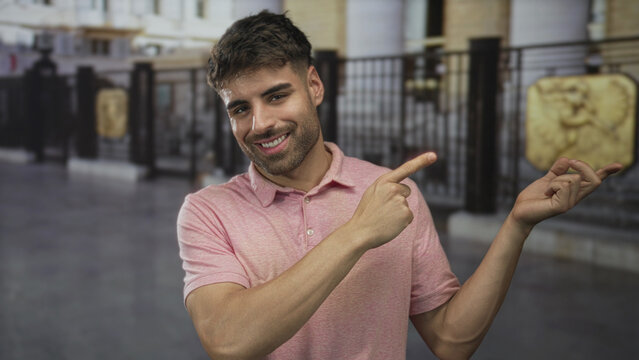 Smiling young man wearing pink shirt pointing finger to iron gate on urban street; optimism hope positivity.