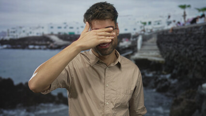 Hispanic man in beige shirt covers his eyes with hand beside a rocky seaside building on stone pier walkway; solitude reflection.