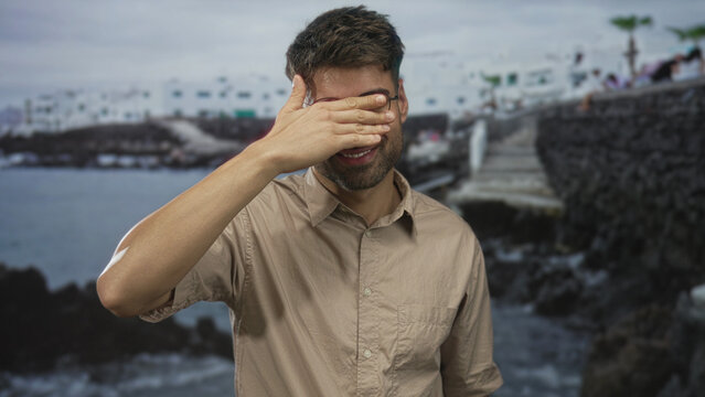 Hispanic man in beige shirt covers his eyes with hand beside a rocky seaside building on stone pier walkway; solitude reflection. - Powered by Adobe