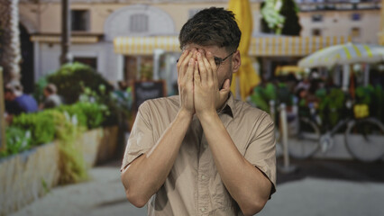 Young hispanic man wearing beige shirt covers face with hands on sunlit city street by cafe seating and bicycles; embarrassment.