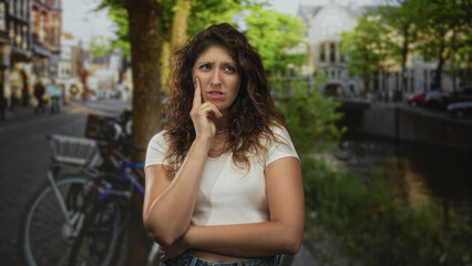 Young hispanic woman with hand on chin and crossed arm standing near parked bicycles by a canal on a city street in daylight; contemplation uncertainty.