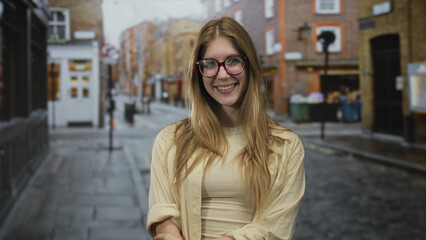 Fototapeta premium Woman with blonde hair and eyeglasses covers her eyes with hands while standing on wet urban street; playfulness.