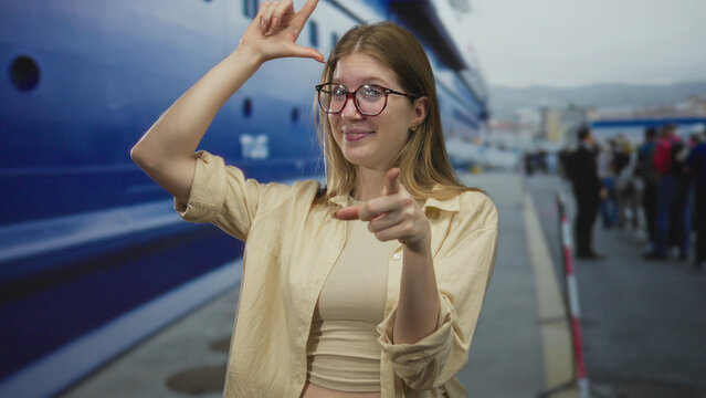 Woman gestures loser sign on forehead near a docked boat and waiting travelers on street by seaside quay; playful.