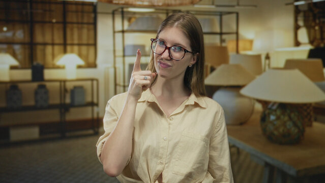 Young blonde woman wearing glasses and a beige shirt shows middle finger beside modern lamp display in a spacious building; defiance anger rejection contempt.