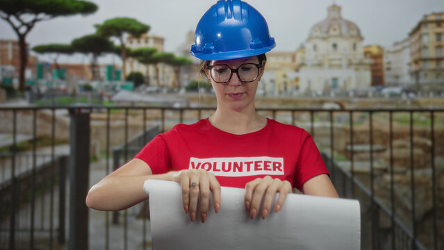 Young blonde woman volunteer in blue hardhat and glasses unrolls white blueprint with bare hands at building near roman ruins; dedication.