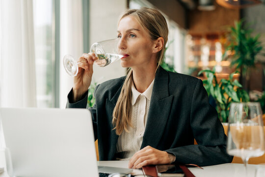 Attractive blond business woman in a modern restaurant drinking water and using laptop for remote work.