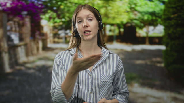 Woman speaking into microphone headset with hand gesture amid lush tree foliage and sunlit green pathway in forest; confidence. - Powered by Adobe