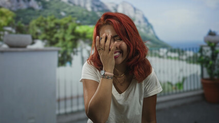 Obraz premium Woman laughing with hand covering face on building balcony wearing white t shirt, red hair and watch, rings visible; joyful.