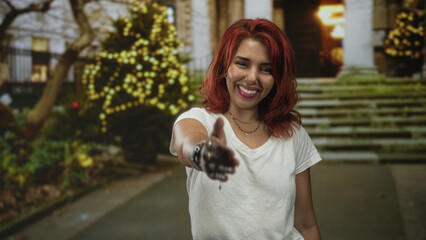 Woman young hispanic extends hand for handshake on urban street steps by stone building entrance with string lights and tree;welcoming greeting.