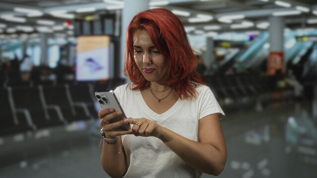 Woman with red hair tapping a smartphone screen with index finger while holding phone in both hands in airport terminal; focused.