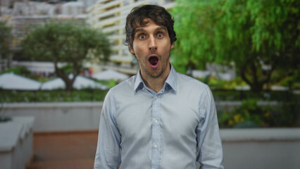 Fototapeta premium Young man with bare face in a light blue shirt glances sideways in a building-lined urban park; serenity.