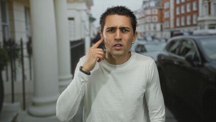 Obraz premium Young hispanic man in white long sleeve shirt holds finger to ear on bustling urban street with parked cars; curiosity.