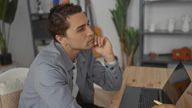 Young man thinking while using a laptop in cozy living room home setting, showing a thoughtful expression alongside guitar and plants.