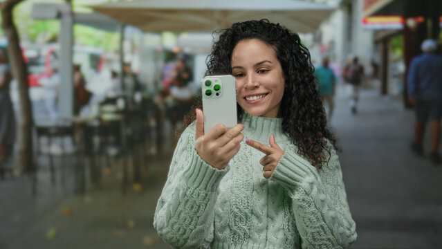 Woman smiling taking selfie with smartphone on terrace in outdoor restaurant setting, displaying happiness and modern technology in lively urban street environment.