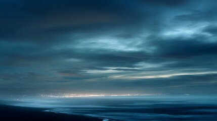 Fototapeta premium Distant city lights glow across a misty ocean horizon under a dark cloudy twilight sky