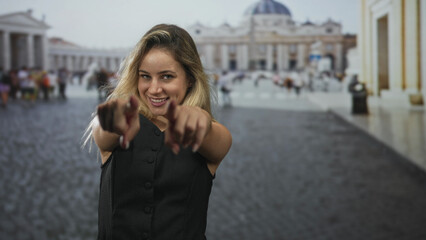 Obraz premium Woman points finger at camera with both hands in square at the vatican, smiling and leaning forward; playful confidence.