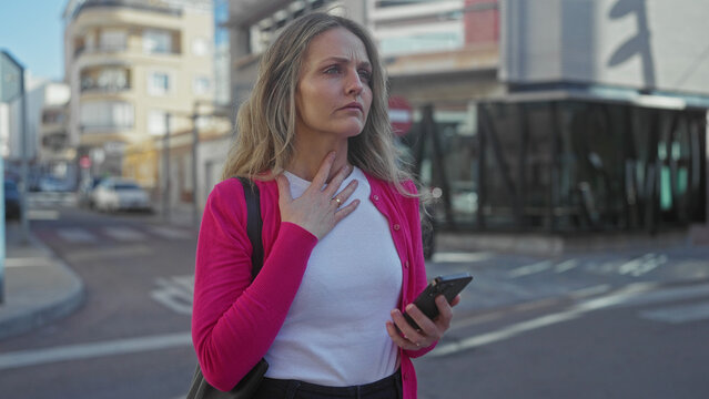 Woman using smartphone outside on urban street with thoughtful expression, wearing pink jacket and white shirt, in modern city setting, young professional appearance, contemporary lifestyle.
