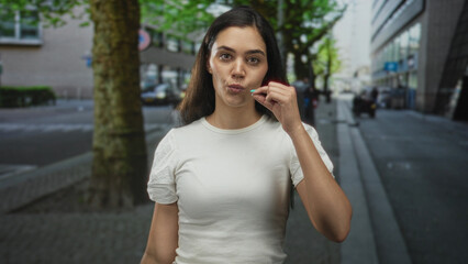 Young hispanic woman covering her mouth with hand on city street, arm raised as if hailing a taxi; surprise concern.