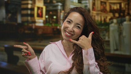 Woman showing call me hand gesture and pointing finger inside ornate church interior; joyful...