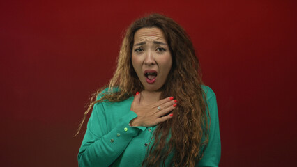 Woman clutching chest with hand and open mouth in red studio wearing green sweater and ring; surprised apology.