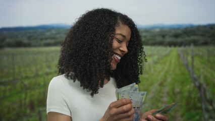 Obraz premium Woman with curly hair smiling and counting banknotes with hands in green forest; prosperity contentment.