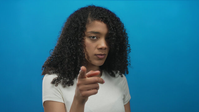 Woman pointing finger in studio with bright blue background, making direct eye contact and raising an index finger near her face; confidence.