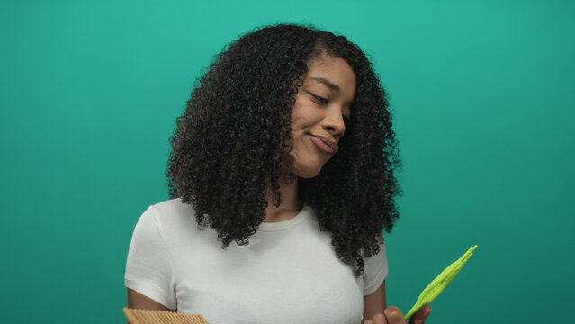 African american woman comparing two combs and checking curly hair texture in studio; choice decision uncertainty.