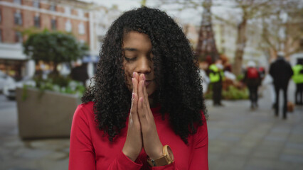 African american woman in red shirt presses palms together on busy urban street; hopeful reflection.