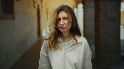 Fototapeta premium Young woman in a white sweater stands inside an old university hallway with arches, expressing a puzzled emotion in a historical setting.