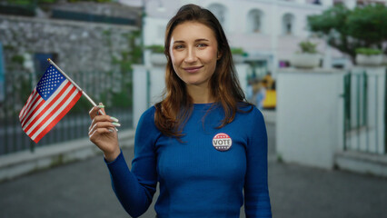 Woman outdoors in city holding american flag and vote badge on blue sweater, symbolizing civic engagement and pride in the united states.