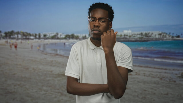 Young man wearing white polo shirt with hand on chin gesture at sunlit coastal building; self reflection calm.