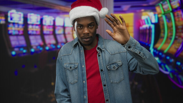 Man waves hand at slot machine in casino building wearing santa hat and denim jacket under neon lights; celebration.