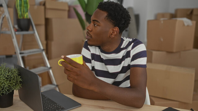 Man holds yellow mug and laptop in building interior, surrounded by unpacked cardboard boxes and a ladder; contentment.