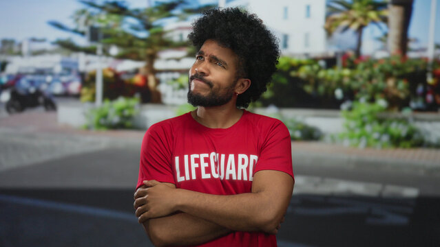 Lifeguard man in red shirt stands with thoughtful expression on a sunny city street with cars and greenery, capturing an engaging urban outdoor scene. - Powered by Adobe