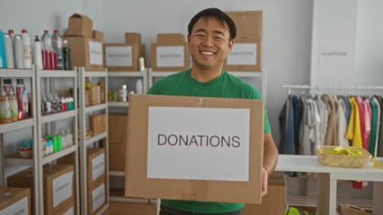 Smiling man in a donation center holding a box labeled donations surrounded by shelves and clothing...