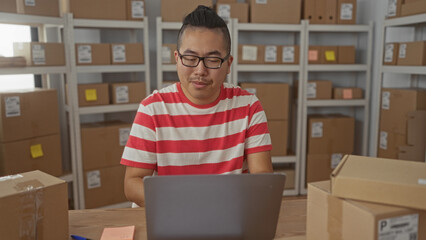 Man organizing parcels and working on laptop at packing table in building office; small business...