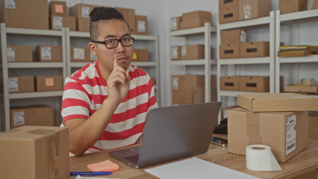 Man typing on laptop and points finger to laptop inside building filled with stacked parcels and shelves while checking labels and tape; focus productivity.