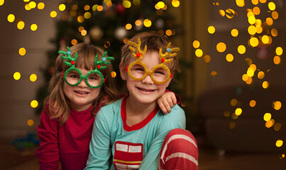 Two kids brother and sister Wearing carnival glasses with Christmas decor on near the Christmas tree at home. 