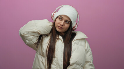 Woman wearing headphones and white winter jacket against a pink background appears thoughtful and stylish, showcasing modern fashion and leisure.