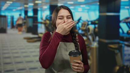 Waitress in apron laughs holding coffee in a bustling gym facility, blending casual service with vibrant sports atmosphere.