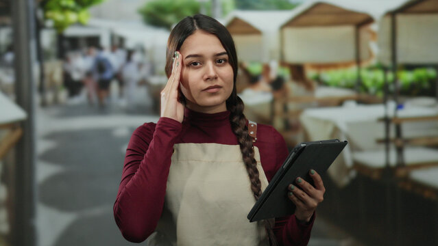 Woman in apron with tablet appears stressed outdoors at a busy restaurant terrace, suggesting her role as a waitress managing orders in a vibrant street setting.
