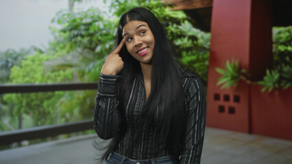 Latin woman in hotel building gestures with finger to head while standing indoors with a playful grimace; craziness.