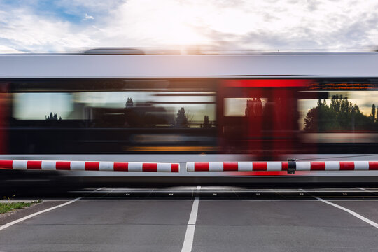 Fast modern regional commute train rushes past closed railway crossing under cloudy sky  sunlight sunset evening. Motion blur rush hour speed dynamic atmosphere  urban transportation commute scene