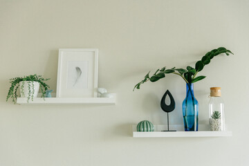 Two white floating shelves on a neutral wall, displaying minimalist home decoration items: framed art, various small plants, sculptures, and a blue glass vase