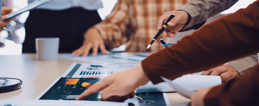 A group of diverse business professionals engaging in a collaborative meeting, analyzing charts and documents on a table in a vibrant office atmosphere. SACTR