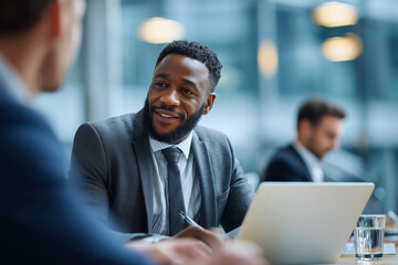 A man in a suit is smiling at another man while they sit at a table with laptops